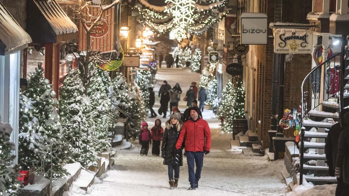 Plusieurs personnes se promènent en soirée sur la rue du Petit-Champlain enneigée et décorée de nombreux sapins illuminés. 