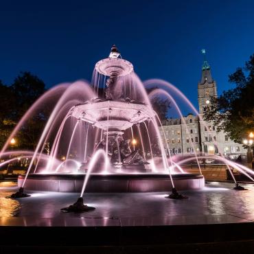 View of the Tourny Fountain illuminated in the evening and the Parliament Building in the background.